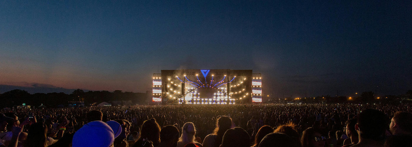 Wide view of an outdoor concert stage at dusk with bright lights and a large crowd