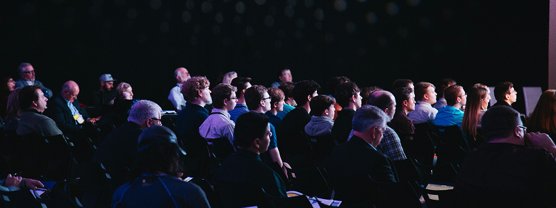 Audience seated in a dark conference hall listening to a speaker
